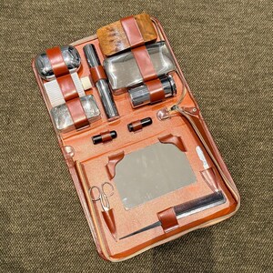 May include: An open, orange leather travel kit with a zipper closure, containing various grooming tools. The kit includes a mirror, comb, scissors, and several silver-toned containers secured with brown leather straps. The kit is designed for travel and personal grooming.