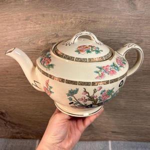 May include: A white teapot with a floral design in pink, green, and gold. The teapot has a round body and a lid with a knob. The teapot is sitting on a wooden surface.