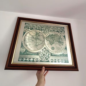 May include: A framed vintage world map print. The map is in shades of green and white, with detailed illustrations of continents and oceans. The frame is a dark brown wood with a gold inner border. The print is held by a hand.