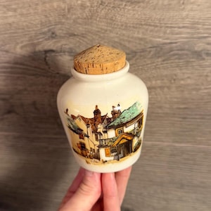Puede incluir: Un pequeño tarro de cerámica de color crema con cuerpo redondeado y cuello estrecho, rematado con un tapón de corcho marrón natural. La parte frontal del tarro presenta una ilustración detallada y colorida de un edificio tradicional con tejado.