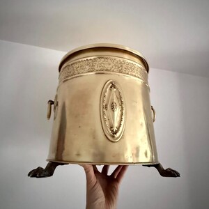 Op de afbeelding: Een vintage messing emmer met een deksel, met decoratieve ovale accenten en een gestructureerde band rond de bovenkant. De emmer heeft twee handvatten en staat op drie klauwpoten. Het messing heeft een warme, gouden tint.
