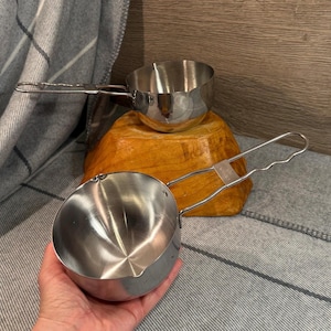 May include: Two stainless steel measuring cups with long handles. One cup is held in a hand, while the other is on a wooden surface. The cups have a shiny, reflective surface, and are designed for kitchen use.