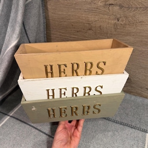 May include: Three wooden herb boxes in natural, white, and sage green. Each box has the word "HERBS" in gold lettering. The boxes are stacked, with the natural wood box on top.
