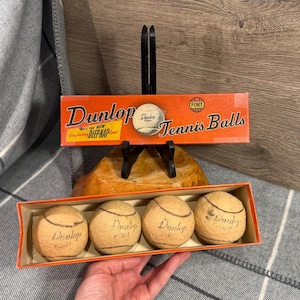 May include: Vintage Dunlop Fort tennis balls in a cardboard box. The box is orange with black text that reads "Dunlop Tennis Balls." The tennis balls are yellow with black text that reads "Dunlop Fort."