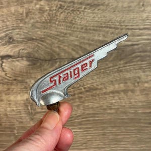 Pode incluir: Um ornamento de capô de carro prateado em forma de asa com a palavra "Staiger" em letras vermelhas.