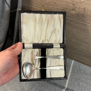 Puede incluir: Un juego de cuchara y utensilio de servir de plata vintage en un estuche negro con forro de satén color crema. El estuche rectangular tiene un pequeño emblema dorado. La cuchara tiene un cuenco redondeado y el utensilio de servir tiene una hoja plana y en ángulo.