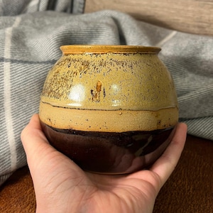 Puede incluir: Un jarrón redondo de cerámica con un esmalte marrón y beige. El jarrón tiene una boca ancha y un fondo redondeado.