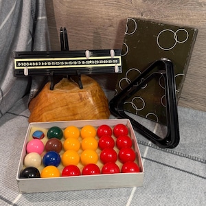 Puede incluir: Un juego de billar vintage con una caja de bolas de colores, un triángulo negro, un dispositivo de puntuación y una caja con un diseño circular. Las bolas son de varios colores, incluyendo rojo, amarillo y negro.