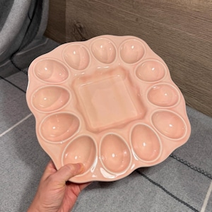 May include: A light pink ceramic egg serving platter with a scalloped edge and a square centre compartment. The tray features twelve oval-shaped indentations arranged around the perimeter, designed to hold individual eggs securely in place.