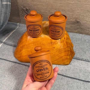 May include: Three terracotta spice jars with lids, labeled "Bay Leaves," "Rosemary," and "Chives." The jars are cylindrical with a rounded top and a small knob. The labels are in a circular design with text. The jars are on a wooden surface.