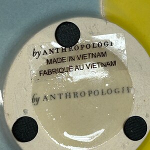 Op de afbeelding: Een witte keramische basis met een zwarte sticker waarop staat "by Anthropologi, Made in Vietnam, Fabriqu&eacute; au Vietnam". De basis heeft drie zwarte stippen aan de onderkant.