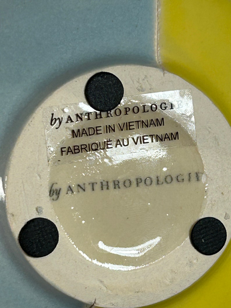 Op de afbeelding: Een witte keramische basis met een zwarte sticker waarop staat "by Anthropologi, Made in Vietnam, Fabriqu&eacute; au Vietnam". De basis heeft drie zwarte stippen aan de onderkant.