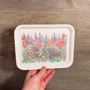 May include: A rectangular white serving tray with a floral design. The tray features a colourful garden scene with red poppies, pink and purple flowers, and green foliage. The tray is held by a hand.
