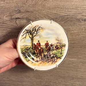 May include: A decorative plate featuring a hunting scene with riders on horseback, hounds, and a landscape with trees. The plate has a gold rim and is held by a white plate hanger.