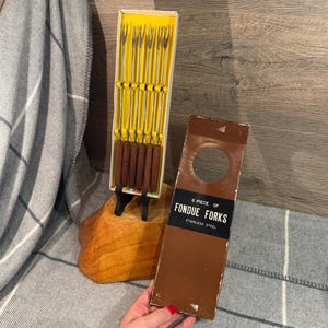 May include: A vintage set of six stainless steel fondue forks with wooden handles, displayed in a yellow and white box. The box is next to a brown cardboard sleeve with the text "6 Piece of Fondue Forks".
