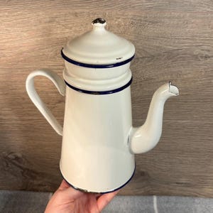 May include: White enamel coffee pot with a blue trim and a lid. The pot has a handle and a spout.