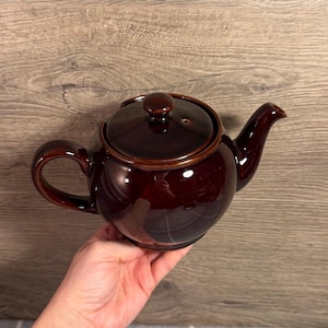 May include: A glossy dark brown ceramic teapot featuring a rounded body, a curved handle, a short spout, and a matching lid with a small knob handle. The surface of the teapot has a reflective finish that shows light highlights across its curved form.