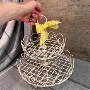 Könnte beinhalten: Ein zweistufiges Serviertablett aus weißem Metall mit dekorativem Drahtdesign. Das Tablett hat einen Griff und ist mit einer gelb-weiß karierten Schleife verziert. Das Tablett ist für die Präsentation und das Servieren von Speisen konzipiert.