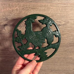 May include: A dark green, circular trivet with a rooster design. The rooster is perched on a branch with leaves, within a circular frame. The trivet is made of cast iron and is approximately 18 cm in diameter.
