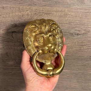 May include: A polished gold-coloured metal door knocker shaped like a lion head. The lion features a detailed mane, prominent eyes, and a nose, with a circular ring held in its mouth. The metal surface reflects light, highlighting the textured features.