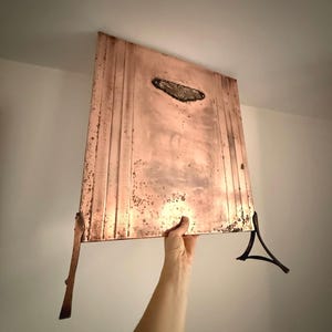 May include: A copper-colored, rectangular wall-mounted light fixture with vertical grooves and a decorative oval accent. The fixture has a distressed finish and is held up by a hand. The fixture has a decorative metal accent.