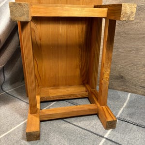 May include: A small, wooden stool with a natural finish. The stool is constructed with square legs and a rectangular top. The wood grain is visible, and the stool appears to be handmade. The stool is shown upside down.