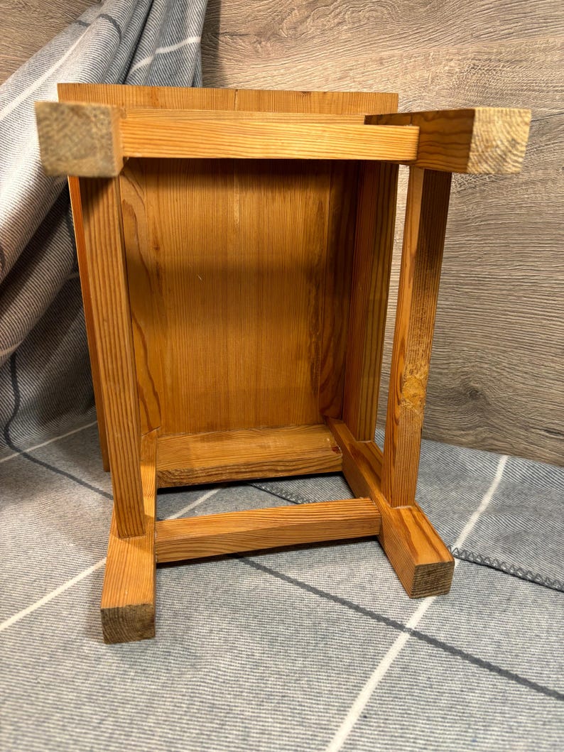 May include: A small, wooden stool with a natural finish. The stool is constructed with square legs and a rectangular top. The wood grain is visible, and the stool appears to be handmade. The stool is shown upside down.