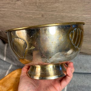 May include: A silver-toned metal bowl with a gold-colored rim and base. The bowl features embossed seashell designs on its sides. The bowl is held by a person.