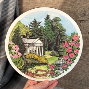 Può includere: Un sottobicchiere rotondo in ceramica bianca con un'illustrazione colorata di un edificio chiamato "The Temple of Flora" a Stourhead, Wiltshire. L'illustrazione rappresenta un edificio bianco con colonne, alberi verdi e fiori rosa.