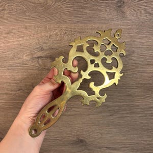 Può includere: Un sottopentola in metallo dorato con un design intricato e decorato. Il sottopentola ha la forma di una croce con un manico lungo.