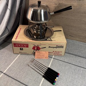 May include: A stainless steel fondue pot with a black handle and lid, sitting on a metal stand. The set includes a box with the text "Fondue Bourguignonne" and several fondue forks with colored tips.