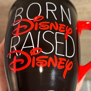 Peut inclure: Mug en c&eacute;ramique noire avec anse et bord rouge. Le mug pr&eacute;sente le texte "BORN DISNEY RAISED DISNEY" en lettres blanches et rouges. Un objet de collection sur le th&egrave;me de Disney.