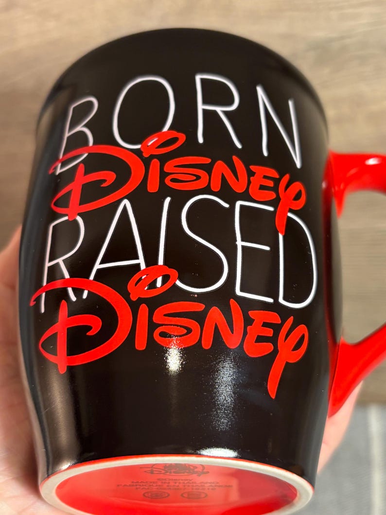 Peut inclure: Mug en c&eacute;ramique noire avec anse et bord rouge. Le mug pr&eacute;sente le texte "BORN DISNEY RAISED DISNEY" en lettres blanches et rouges. Un objet de collection sur le th&egrave;me de Disney.