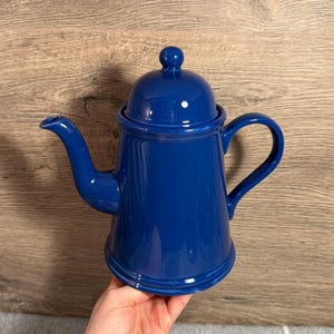 以下が含まれることがあります： コバルトブルーのセラミックコーヒーポット。蓋とカーブしたハンドルが付いています。注ぎ口と丸みを帯びたベースがあります。背景は木製の表面です。