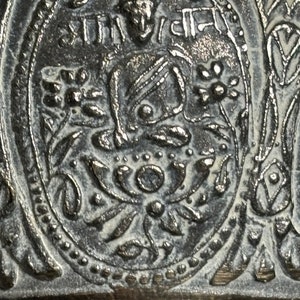 K&ouml;nnte beinhalten: Nahaufnahme einer silbernen Glocke mit aufwendigen floralen Mustern und einem zentralen Bild einer sitzenden Figur. Die Glocke ist mit dem Text "SHTI" in einer dekorativen Schriftart beschriftet.