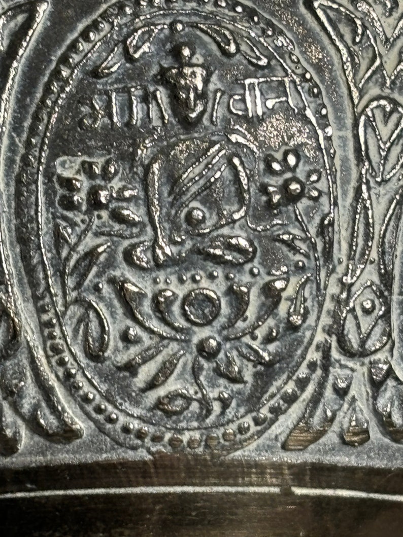 K&ouml;nnte beinhalten: Nahaufnahme einer silbernen Glocke mit aufwendigen floralen Mustern und einem zentralen Bild einer sitzenden Figur. Die Glocke ist mit dem Text "SHTI" in einer dekorativen Schriftart beschriftet.