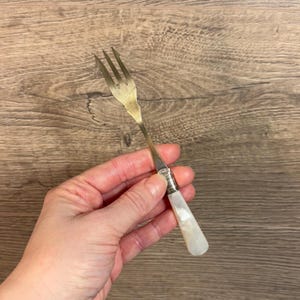 May include: A gold fork with a white mother-of-pearl handle. The fork is held in a hand.