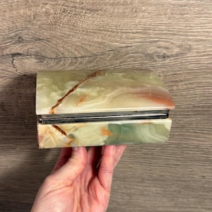 以下が含まれることがあります： 長方形の宝石箱。ヒンジ付きの蓋が付いています。箱は緑と茶色のオニキス製で、茶色の縞模様があります。箱には銀色の金属フレームとヒンジが付いています。