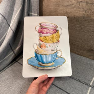 May include: A rectangular white sign featuring a stack of five colorful teacups and a saucer. The teacups are painted in shades of pink, yellow, white, and blue, with gold accents. The teacups are stacked on top of each other, creating a whimsical design.