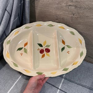 Op de afbeelding: Crèmekleurige, ovale serveerschaal met drie compartimenten. De schaal is versierd met handgeschilderde bladeren in groen, geel en oranje, en trossen rood fruit. De rand heeft een geschulpte rand met een groene rand.