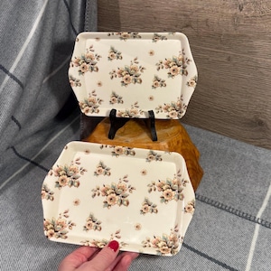 May include: Two rectangular serving trays with a floral design. The trays feature a cream-coloured base adorned with a repeating pattern of pink, peach, and green flowers. They are displayed on a wooden surface.