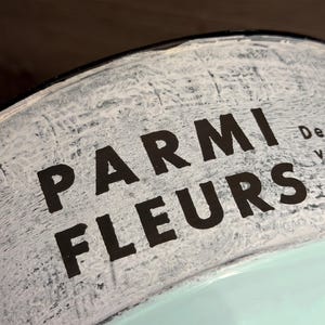 Op de afbeelding: Een gebogen metalen container met een witte, verweerde afwerking en de zwarte tekst PARMI FLEURS op het bovenste gedeelte. Het onderste deel van het object is geschilderd in een effen lichtmuntgroene kleur met een glad en schoon oppervlak.