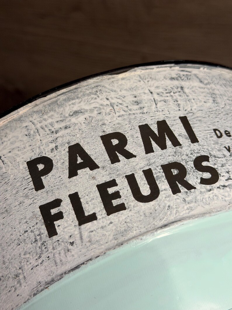 Op de afbeelding: Een gebogen metalen container met een witte, verweerde afwerking en de zwarte tekst PARMI FLEURS op het bovenste gedeelte. Het onderste deel van het object is geschilderd in een effen lichtmuntgroene kleur met een glad en schoon oppervlak.