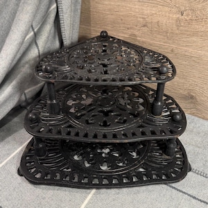 May include: Black, three-tiered corner shelf with an ornate, openwork design. Each shelf is triangular with rounded corners and supported by three posts. The shelf has decorative ball feet and a ball finial at the top.