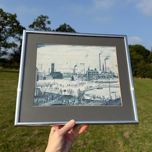 May include: A framed watercolor painting depicting an industrial town scene with factories, a church, and numerous figures. The artwork is framed in a silver metal frame with a gray matting. The painting is held by a person in a grassy outdoor setting.