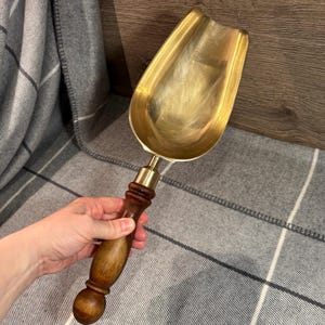 Peut inclure: Une pelle à cheminée en laiton avec un manche en bois. La pelle est de couleur dorée et a une pelle arrondie. Le manche est brun foncé et a un bouton arrondi à l'extrémité.