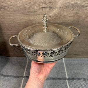 May include: Silver serving dish with a lid and ornate design. The lid has a decorative finial. The dish has a scalloped edge and a cut-out design.
