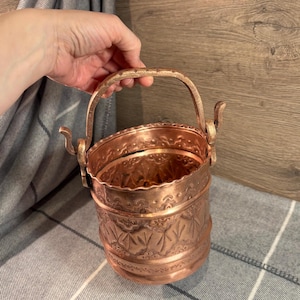 Pode incluir: Um balde de cobre com um design ornamentado e uma alça. O balde é feito de cobre martelado e tem um padrão decorativo ao redor da parte superior e inferior.