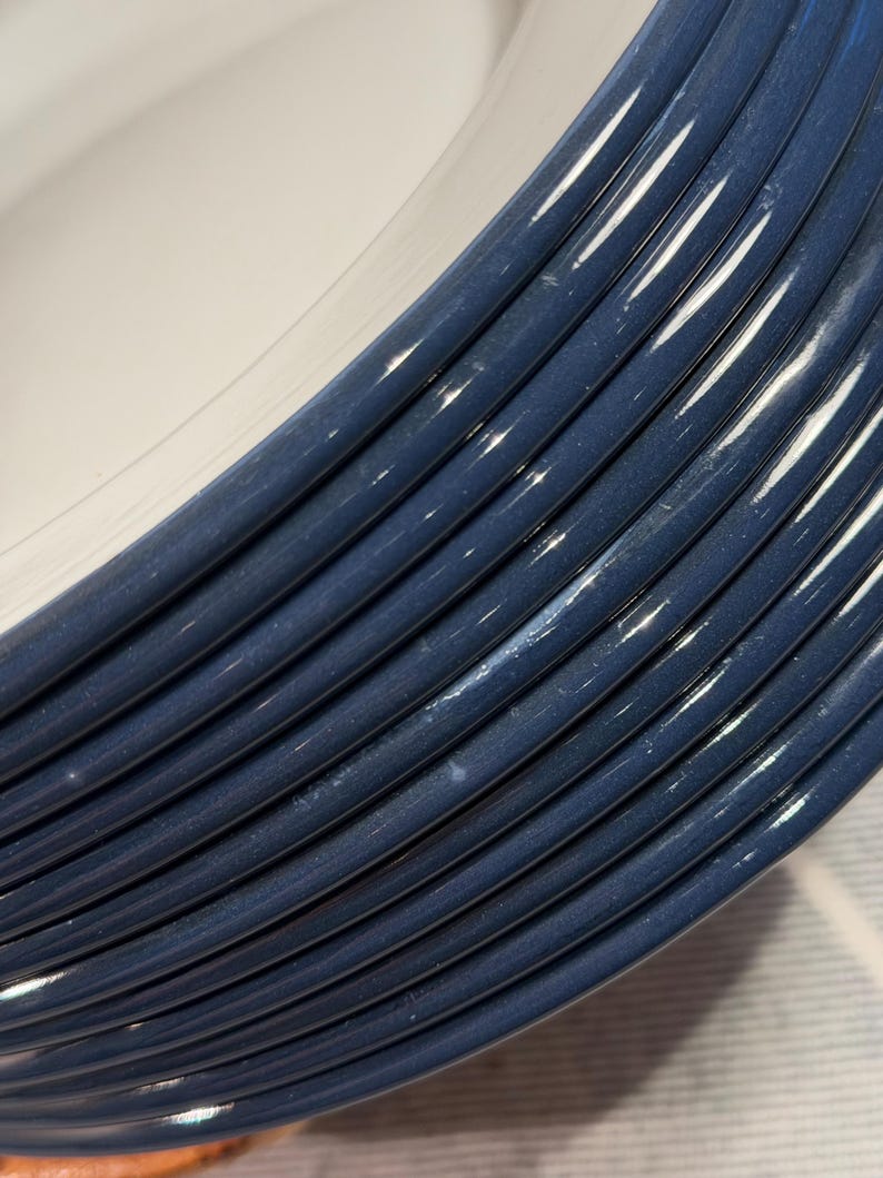 Pode incluir: Uma pilha de pratos redondos de cer&acirc;mica com um centro branco brilhante e uma borda larga azul-escura com uma s&eacute;rie de sulcos circulares conc&ecirc;ntricos em relevo que criam uma apar&ecirc;ncia texturizada e ranhurada na borda de cada prato.