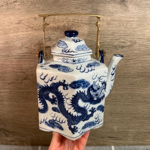 Puede incluir: Tetera de porcelana blanca y azul con diseño de dragón. La tetera tiene forma cuadrada, tapa y asa dorada. El dragón está representado en azul y la tetera tiene una estética asiática tradicional.